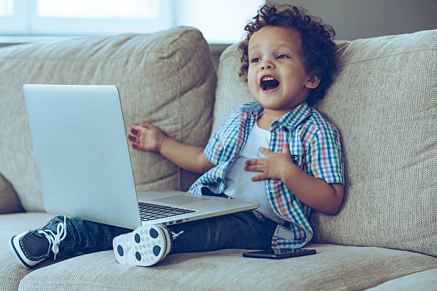 Mom how does this thing turns on? Little African baby boy keeping mouth open while sitting on the couch at home with laptop on his knees creaming stock pictures, royalty-free photos & images