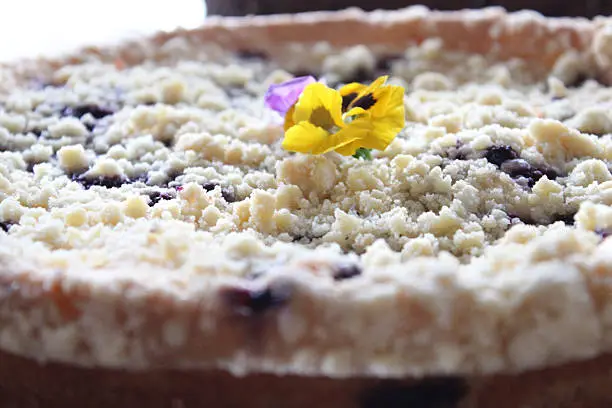 Photo showing a homemade blueberry cooked cheesecake with a crunchy crumble topping, decorated with a yellow edible pansy flower and waiting to be served.