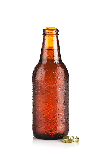 Full brown beer bottle isolated on white background with drops and condensation, back lit and showing a soft white reflection at right side. The bottle is opened and a golden cap is at the base of it.
