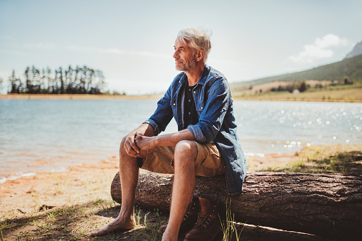 Portrait of a mature man sitting on a log near the lake. Senior caucasian man sitting alone on the lake and looking at a view on a summer day.