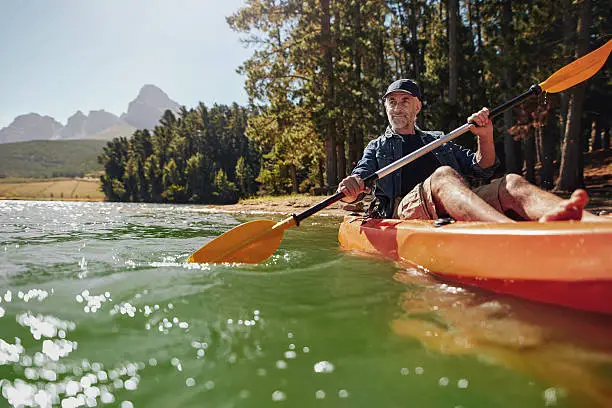 Portrait of a mature man with enjoying kayaking in a lake. Caucasian man wearing a cap paddling a kayak on summer day.