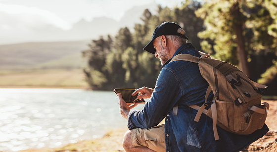 Senior man with backpack using digital tab while sitting near a lake. Mature man on hike in nature using digital tablet.