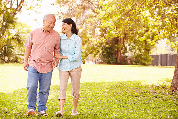 Senior Asian Couple Walking Through Park Together Senior Asian Couple Walking Through Park Together Holding Hands Smiling locking-arms stock pictures, royalty-free photos & images