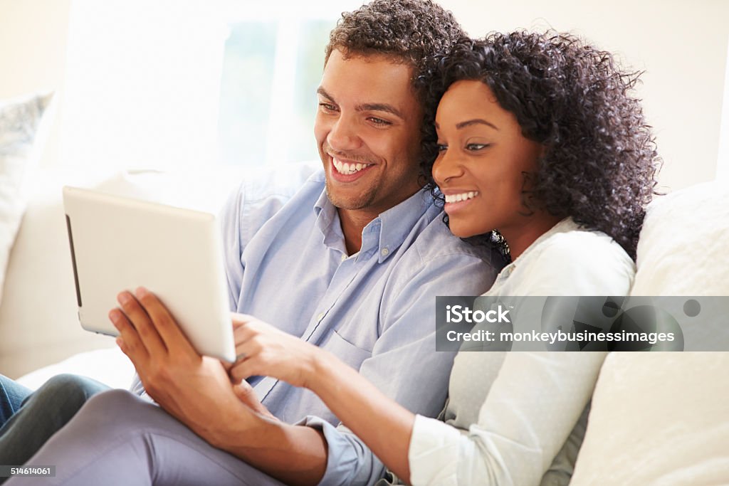 Young Couple Sitting On Sofa Using Digital Tablet Young Couple Sitting On Sofa Using Digital Tablet Smiling Digital Tablet Stock Photo