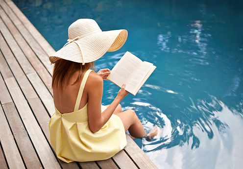 Rearview shot of a woman relaxing by the pool with a book