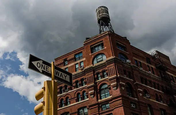 Historic brick building in New York City with water tower on top, stoplight and one way sign in foreground, clouds in blue sky background