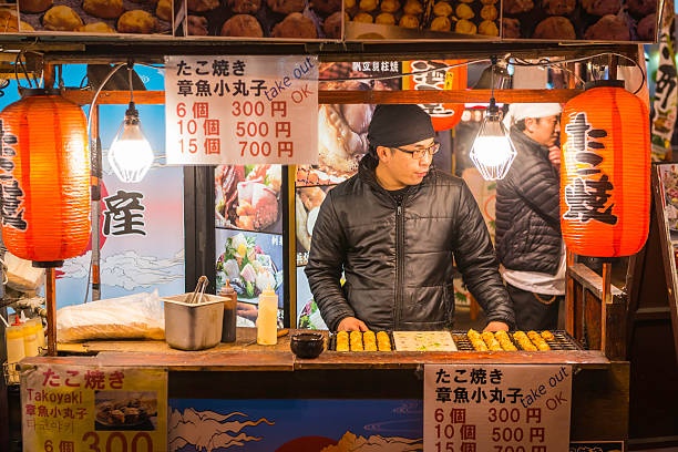 Japan cook selling takoyaki snack food night market stall Osaka Osaka, Japan - February 27, 2016: Cook selling traditional takoyaki, the popular fast food snack of batter and octopus, at a street food market stall in the Dotonbori district of Osaka, Japan's vibrant second city. cooking takoyaki is a ball shaped japanese snack stock pictures, royalty-free photos & images