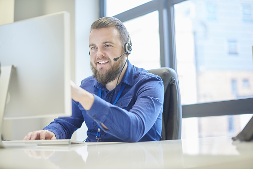 a call centre phone operative in his mid 30s chats on the phone at his desk . He looks in complete control of the situation and smiles through the mass of fur .