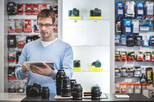 Store clerk is standing inside photographic store and holding digital tablet. On the desk in front of him are digital camera and various lenses.