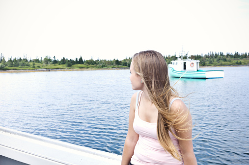 Beautiful Young Woman on a Boat, Mahone Bay Nova Scotia.  She sits on the side deck, ocean behind and good copy space.  Stock photo taken with a Leica M9 camera.