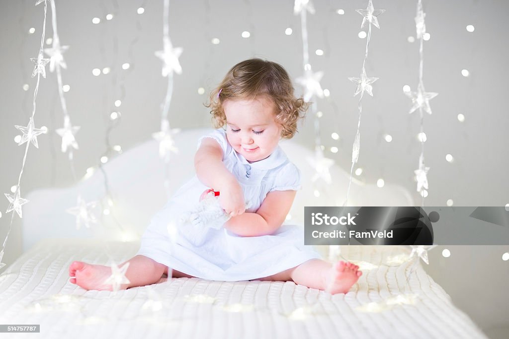 Adorable toddler girl with toy in bed between Christmas lights Adorable toddler girl with curly hair wearing a white dress is playing with a toy in a white bed between Christmas lights Baby - Human Age Stock Photo