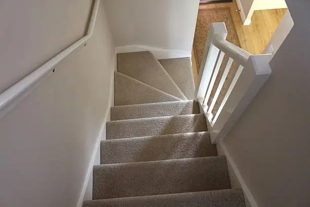 Photo showing the view looking down a beige carpeted stairway with a white, wooden handrail and balustrade. A rug can be seen on the wooden floor of the  entrance hall.