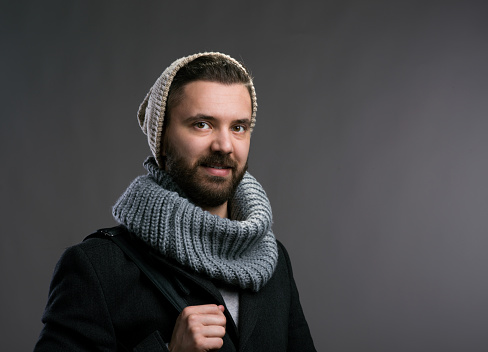 Young hipster man in black winter coat, knitted scarf and beanie. Studio shot on gray background.