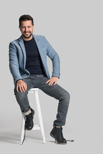 Model in hat and glasses is sitting on a chair. Handsome man is in the studio against white background.