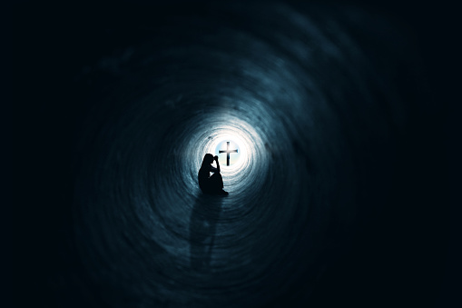 A woman and her shadow praying in the darkness of a tunnel with a cross and bright light just on the outside.