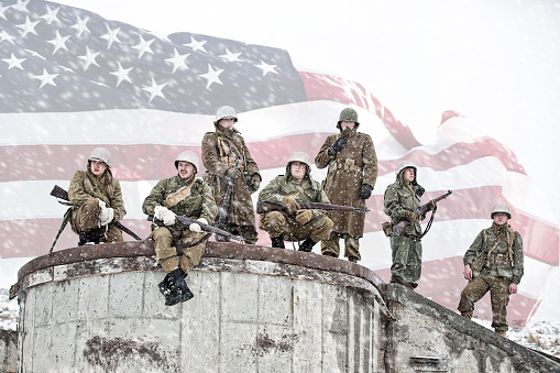 Platoon of American WWII soldiers resting on top of the foundation of a bombed out building. American flag overlay.