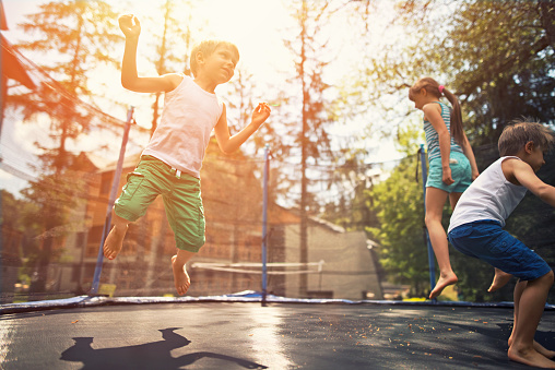 Little boys aged 5 and the girl aged 9 having fun on garden trampoline. Evening sun shining brightly. 