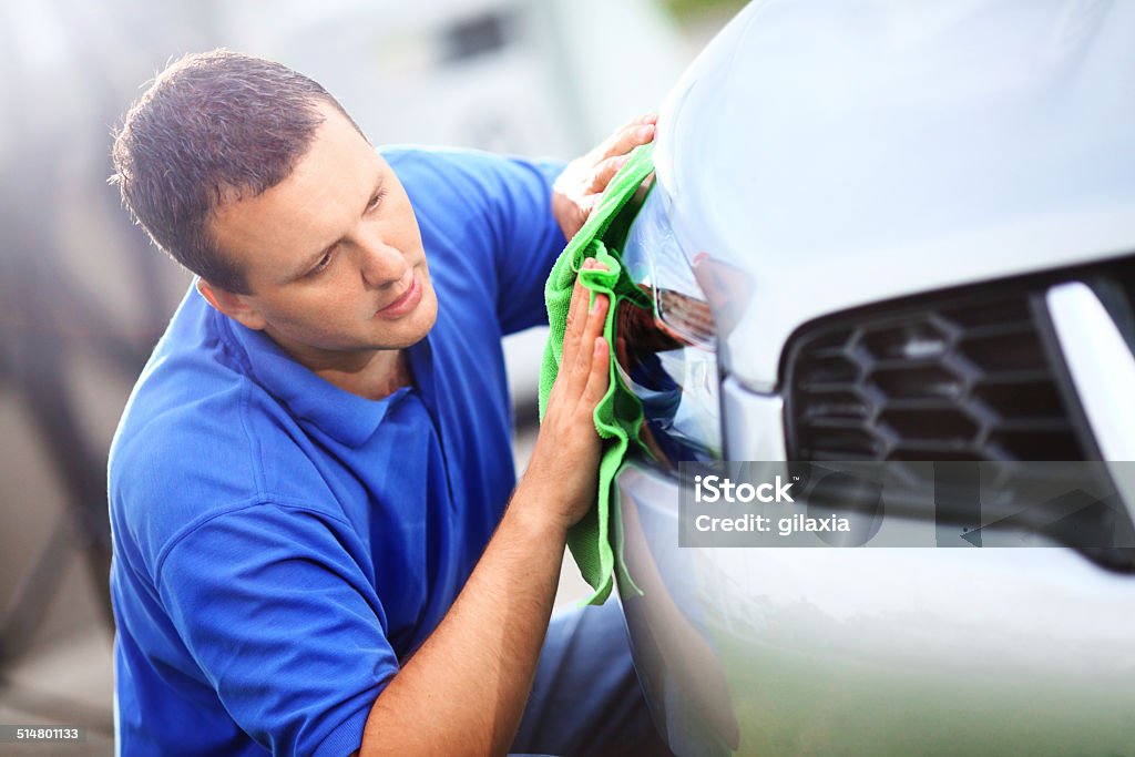 Car wash. Mid-aged man polishing headlight on his silver metallic colored vehicle with green cloth at parking lot. He's kneeling next to his car.He has brown hair and wearing blue polo shirt. Cleaning Stock Photo