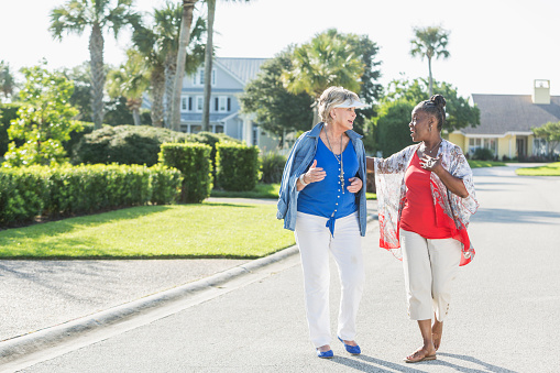 Two multi-racial senior woman, one Caucasian and the other African American, taking a walk in a residential neighborhood on a bright, sunny summer or spring day. They are enjoying each other's company, smiling, face to face.