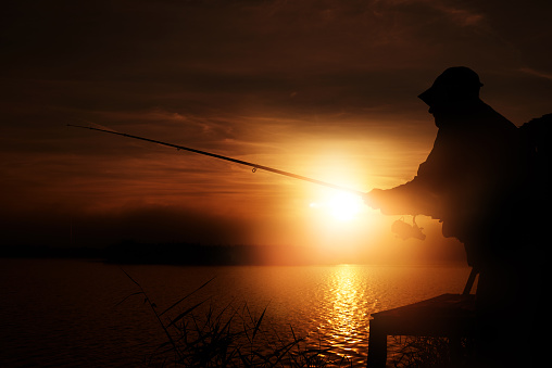 In silhouette of adult fisherman standing by the river and throwing the spinning. Lifestyle. Sport and hobby