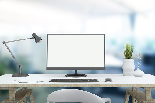 View at a working desk,  with a large PC monitor, a lamp, a plant, with a blurry out of focus business background.