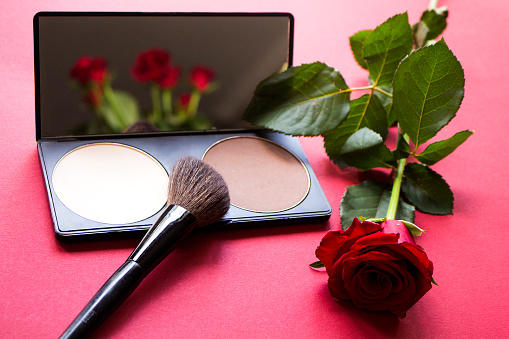 Correcting powder, brush and red rose on dark pink textured surface. Roses reflecting in the mirror. Makeup product to even out skin tone and complexion. Professional cosmetics.