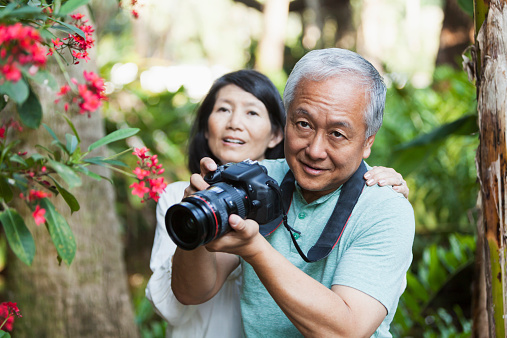 Senior Asian couple (60s) in park taking photographs.  Focus on man.