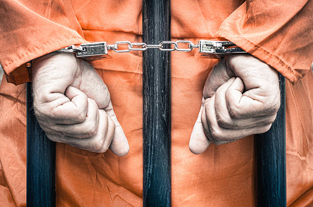 Handcuffed hands of a prisoner behind prison bars Handcuffed hands of a prisoner behind the bars of a prison with orange clothes - Crispy desaturated dramatic filtered look torture photos stock pictures, royalty-free photos & images
