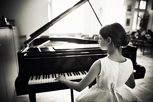 Black and white photo of little girl playing grand piano in a sunny class room. She is preparing for a piano performance in music school. The girl aged 9 is sitting at the grand piano, back to the camera. The girl is wearing a white dress.
