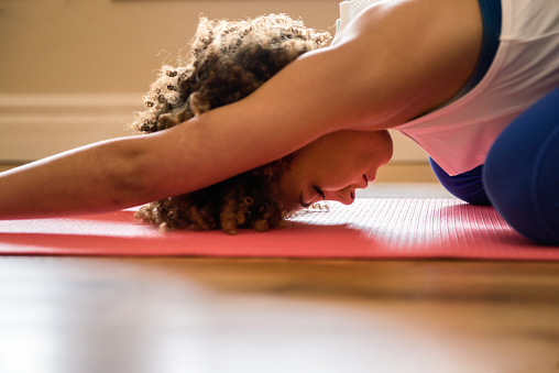 Pretty, athletic young woman practicing Yoga Child's pose on a blue yoga mat in an empty room with a wood floor.  Weights and bottles of water in the background.