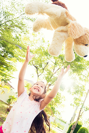 This is a horizontal, color photograph of a happy, 6 year old Thai girl playing in her yard with her stuffed animal. She smiles with missing teeth as she throws her dog in the air. Bright light and green trees fill the background. Focus is soft.