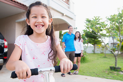 Young family of Thai and American descent spends time in the front yard of their home in Nakhon Ratchasima, Thailand. The parents stand together in the background as their smiling, 6 year old daughter on a scooter fills the foreground.