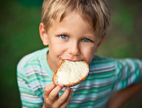 Portrait of little boy aged 5 eating loaf of bread with butter outdoors. Summer vacations.