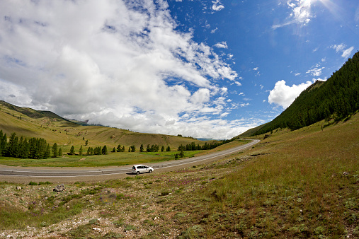 Rural mountainds landscape with hills, mountains, road, blue summer sky with clouds and sun and car parked at the roadside during a trip over Altai region (Russia).