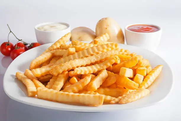 Portion of French fries (Crinkle-cut) deep fried, served on a white plate next to white bowls with mayonnaise and ketchup and fresh potato.