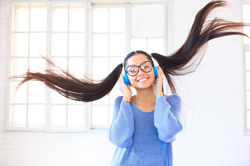Attractive asian young girl with blue headphones, eyeglasses and ponytails, blue blouse, listening music and dancing. On white background with windows.