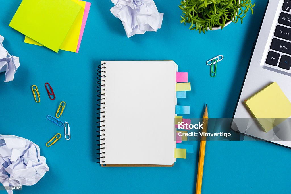 Office table desk with set of colorful supplies, white blank Office table desk with set of colorful supplies, white blank note pad, cup, pen, pc, crumpled paper, flower on blue background. Top view and copy space for text. Advertisement Stock Photo