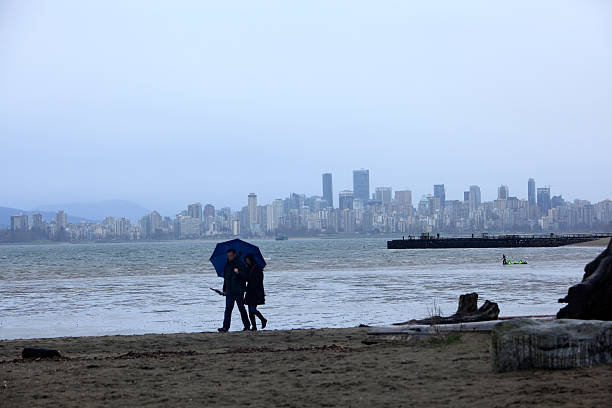 Young Couple Enjoying A  Rainy Walk OnThe Beach. Vancouver,Canada- March 13,2016: Young Heterosexual couple with Umbrella walking a Vancouver Beach in the rain.  English Bay and Vancouver skyline in the background. Pier with people mid frame plus fellow with kite in water.  beach english bay vancouver skyline stock pictures, royalty-free photos & images