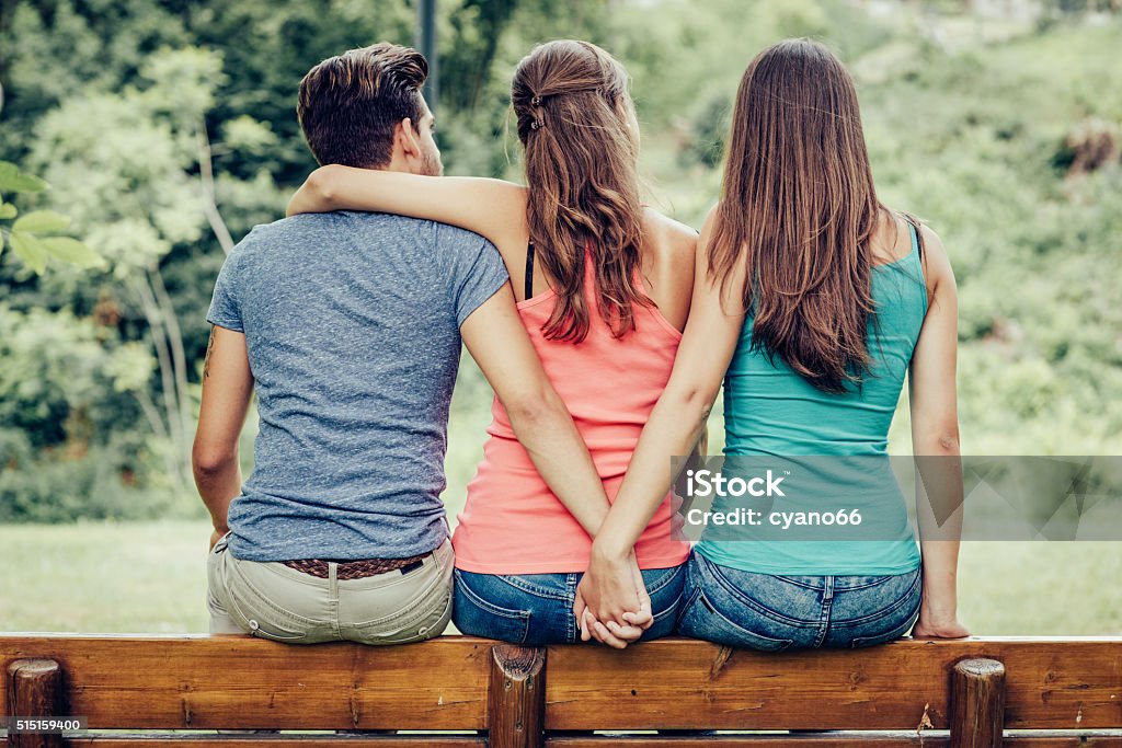 Love triangle Love triangle, a girl is hugging a guy and he is holding hands with another girl, they are sitting together on a bench Infidelity Stock Photo