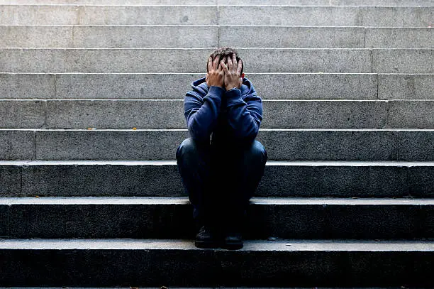 Young desperate man in casual clothes abandoned lost in depression sitting on ground street concrete stairs alone suffering emotional pain, sadness, looking sick in grunge lighting