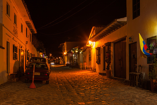 Villa de Leyva, Colombia - September 13, 2014:   The sun has set over Villa de Leyva on the Andes Mountains in Colombia; Calle or Street 14 is now dimly lit with lamp lights. The sky is completely dark. Some restaurants on the street fly the flag of country whose cuisine figures on their menu; a few cars are parked on the old cobblestoned street.  Founded in 1572 and located at just over 7000 feet above sea level on the Andes Mountains, Villa de Leyva was declared a National Monument in 1954 to protect it's colonial architecture and heritage. It is located in the Department of Boyaca, in the South American country of Colombia. The town is a popular conference destination.     Photo shot at night; a four second exposure causes motion blur where there are people or flags moving. Horizontal format; copy space.