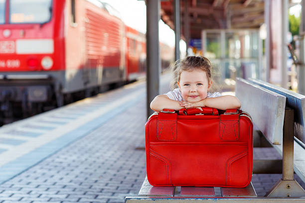 Funny little girl with big red suitcase on a railway station. Kid waiting for train and happy about traveling.