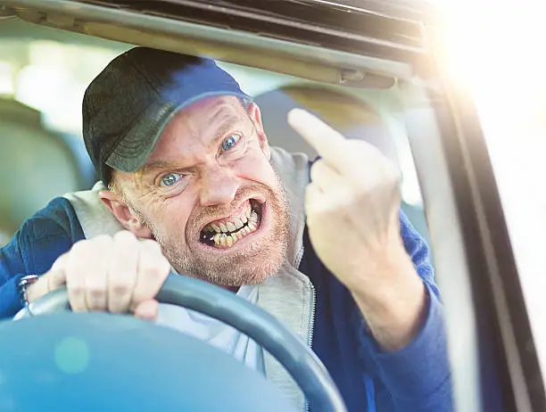 Losing control, a furiously angry male driver makes an obscene gesture, his teeth clenched,  through the car windshield in a bout of uncontrollable road rage!