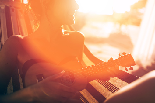 Smiling girl enjoys her music, playing an ukulele while sitting in a hammock on the porch on a sunny summer day 