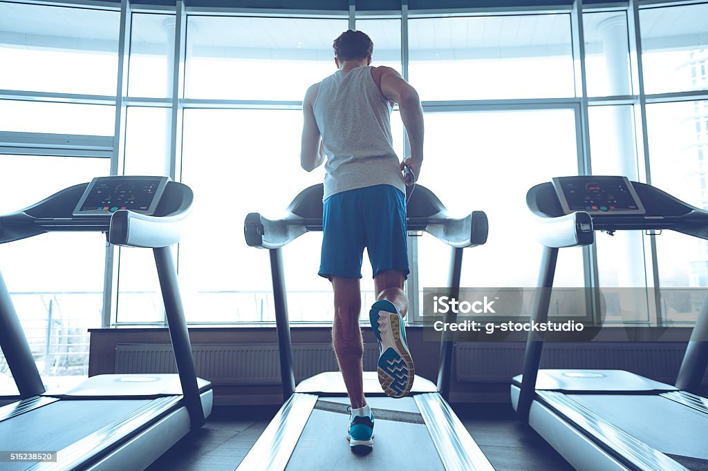 Jogging his way to good health. Full-length rear view of young man in sportswear running on treadmill in front of window at gym Treadmill Stock Photo