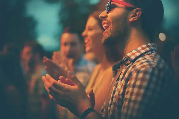 Group of people clapping and cheering after music performance. Standing side by side. There are two guys and one girl and large crowd behind.The closest guy is in focus.Side view.