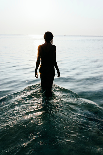 Tranquil scene with a view from behind of the silhouette of a wet young woman standing in the ocean facing out in meditation as the water flows around her legs creating a rippling pattern