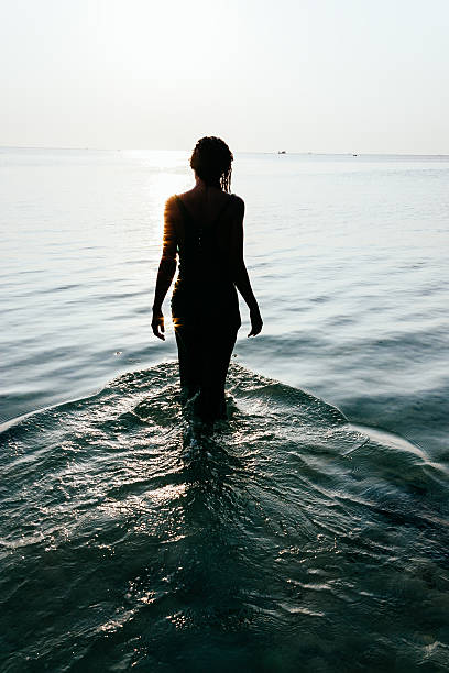 Silhouette of a wet woman standing in the ocean Tranquil scene with a view from behind of the silhouette of a wet young woman standing in the ocean facing out in meditation as the water flows around her legs creating a rippling pattern wading stock pictures, royalty-free photos & images