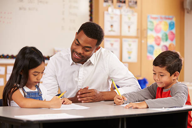 istock Teacher sitting at desk with two elementary school pupils 515262088