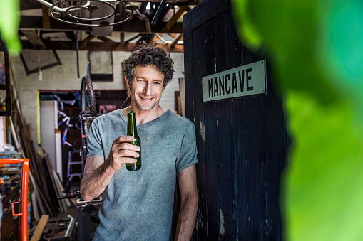 A photo of happy worker holding beer bottle at doorway. Portrait of craftsperson is in casuals. Confident repairman is in his workshop.